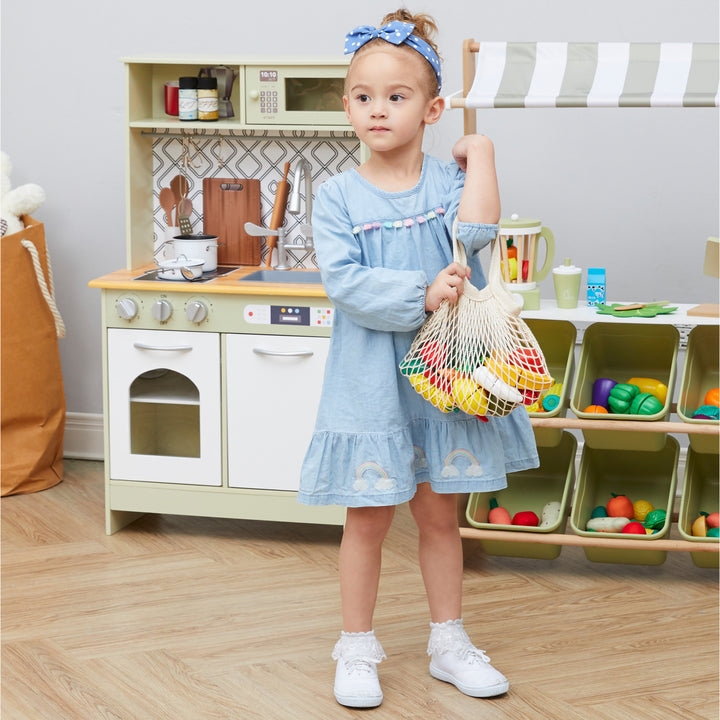 A young girl holding a Teamson Kids Little Chef Frankfurt 21 Piece Wooden Produce Shopping Bag Set with Pretend Fruit and Vegetables, Multicolor, standing in front of a play kitchen playset with kid-sized dimensions.