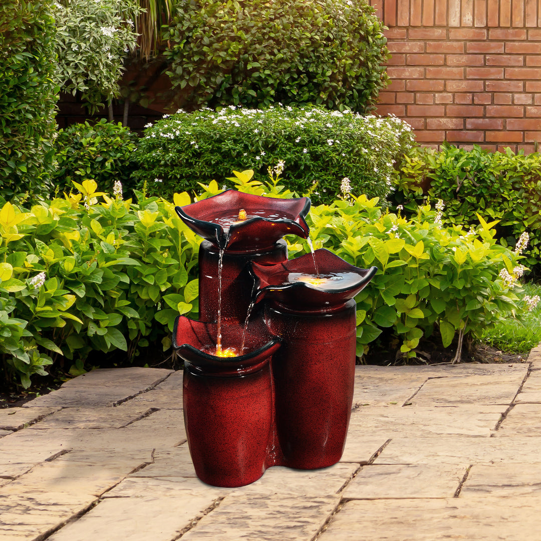 A Teamson Home outdoor water fountain with a rustic red finish set against a garden backdrop, featuring LED lights for enchanting garden decor.