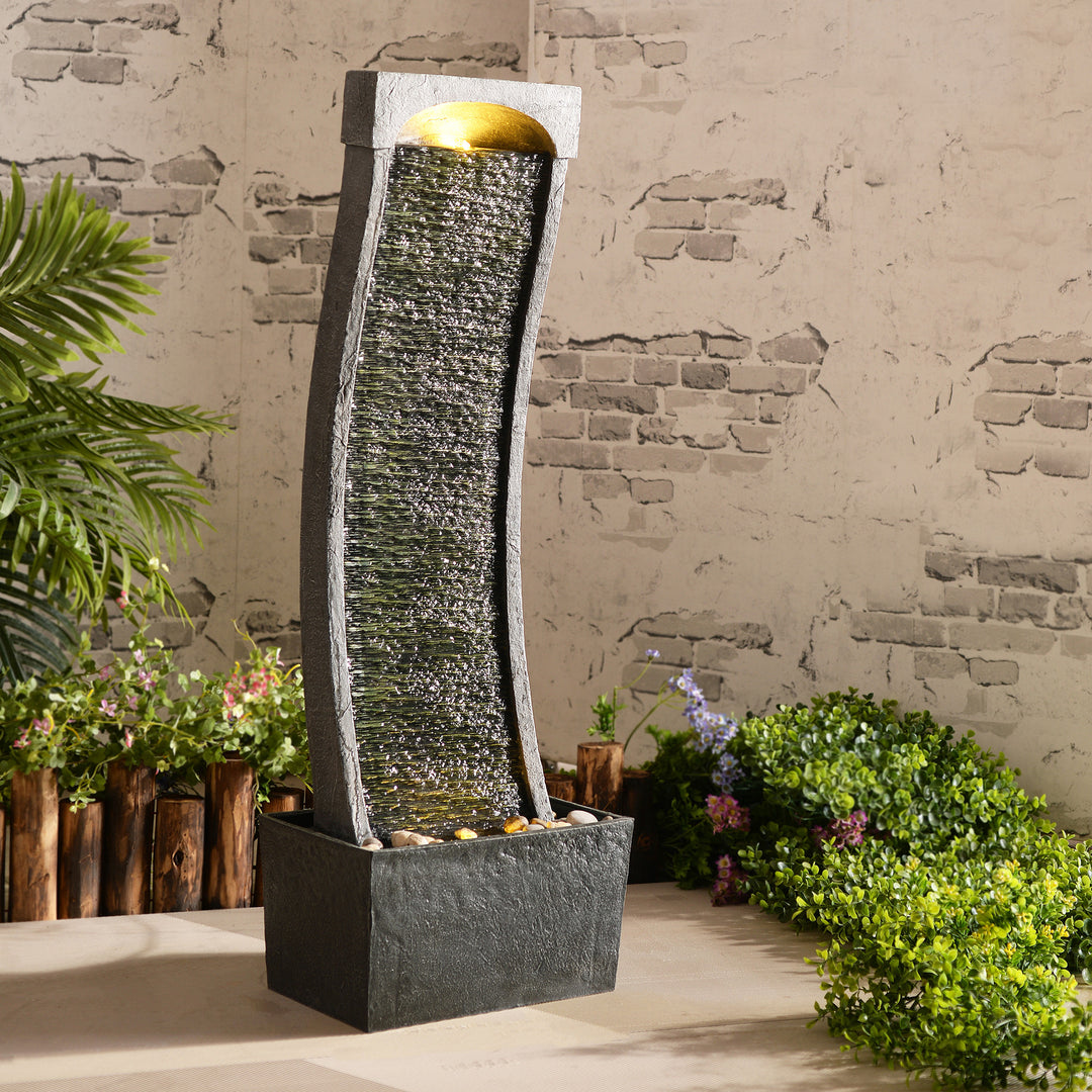 Teamson Home Indoor/Outdoor Modern Curved Slate Waterfall Fountain with LED Lights in an outdoor setting with shrubs and flowers