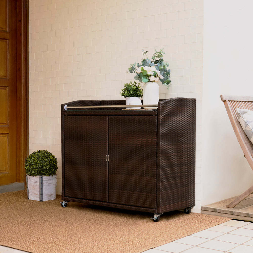 Teamson Home Veronica Portable Brown PE Rattan Outdoor Bar Carton wheels with a vase and plant on top, placed in a home entrance area.