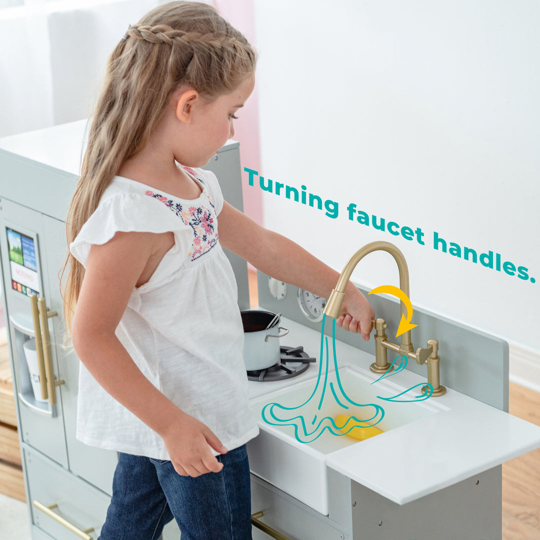 A young girl turning the handles of a Teamson Kids Little Chef Charlotte Modern Play Kitchen, Silver Gray/Gold sink faucet part of her playset kitchen.