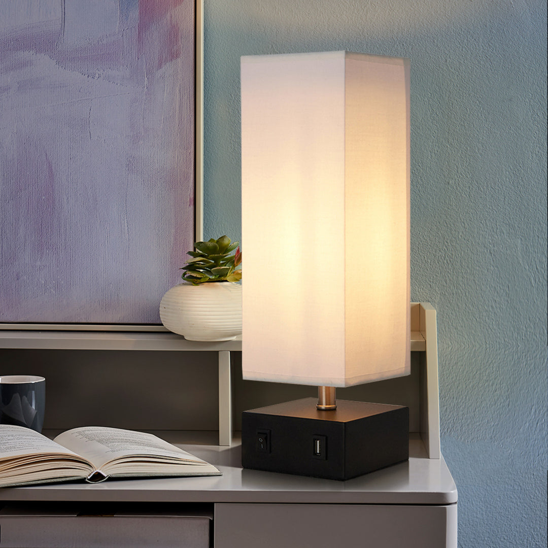 A Teamson Home Colette 14.5" Modern Metal Table Lamp with Square Shade and USB Port in Black/White on a desk next to a book.