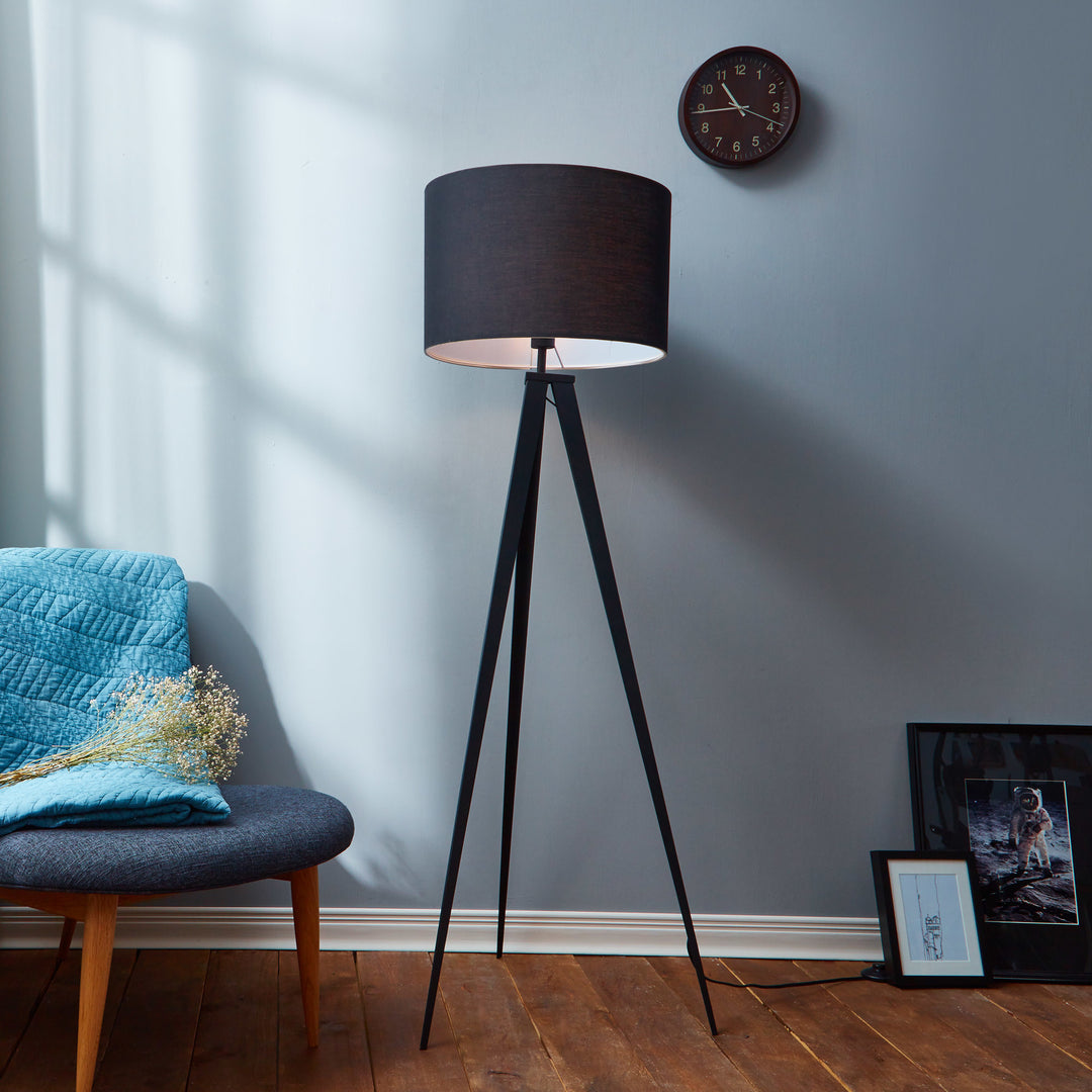 A versatile Teamson Home Romanza 60" Postmodern Tripod Floor Lamp with Drum Shade, Matte Black stands next to a blue armchair with a quilted throw pillow, against a gray wall with light streaming through blinds, accompanied by a wall clock and picture frames.