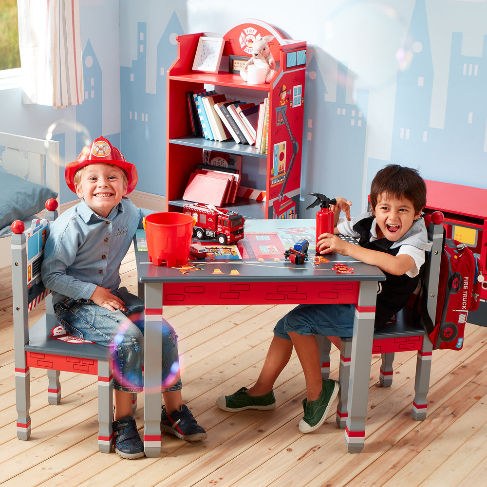 A boy with blonde hair and a pretend firefighters' helmet and a boy with brown hair and a pretend fire fire extinguisher sit across from each other at a child's fireman themed table in a firemen themed room.