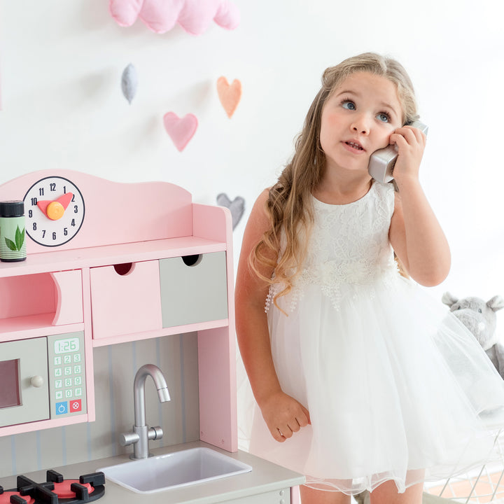 A young girl playing pretend on a toy phone next to the Teamson Kids Little Chef Florence Classic Play Kitchen, Pink/Gray.