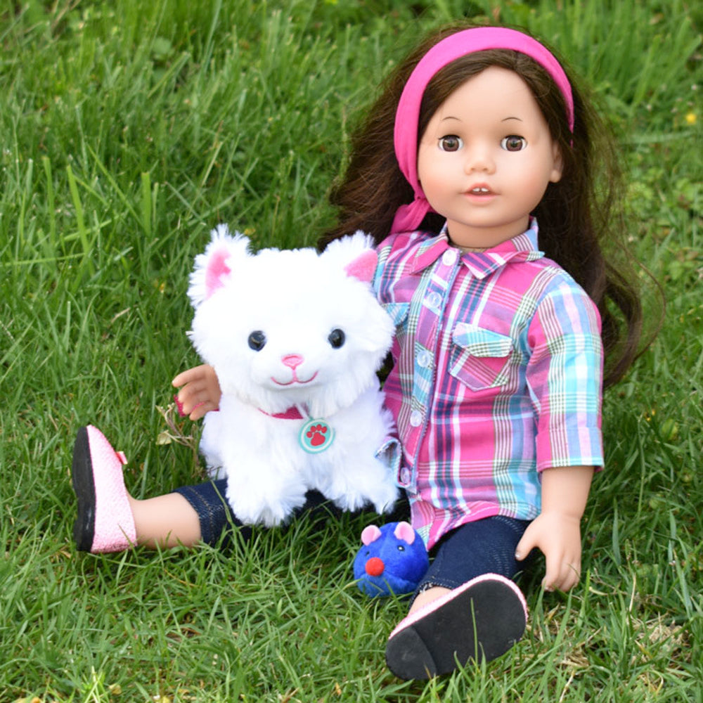 A brunette 18" doll with a plaid shirt and denim pants on sits in the grass with her faux white kitten companion.