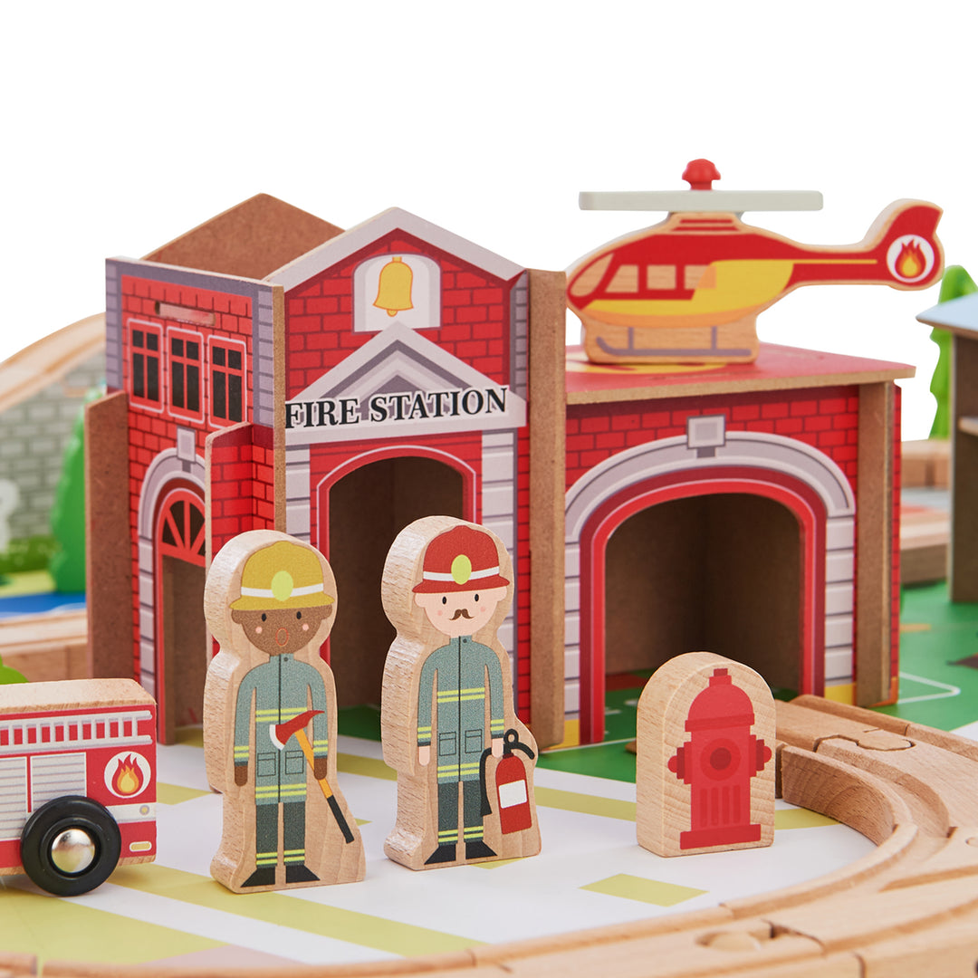 A illustrated fire station next to the train tracks with a helicopter on the roof, a fire hydrant, two firefighter figurines, and a fire truck.