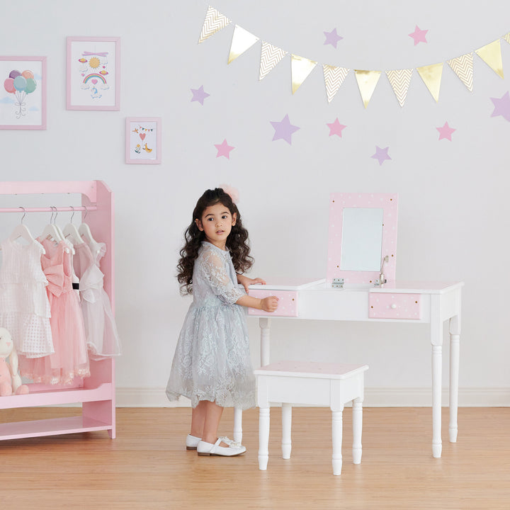 A little girl standing in front of a Fantasy Fields Kids Kate Twinkle Star Vanity Set with Foldable Mirror and Chair, Pink/White.