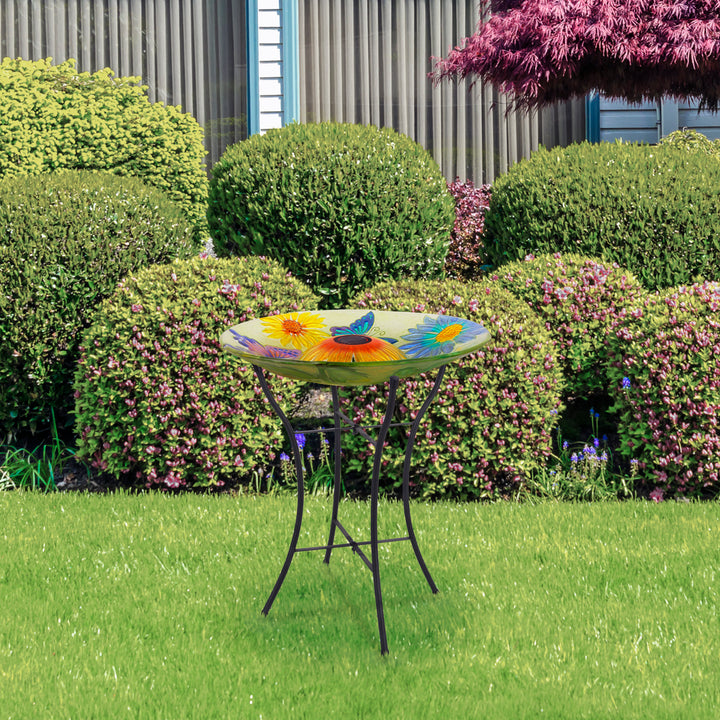 A colorful, flower-patterned Teamson Home Outdoor 18" Hand painted Butterfly Fusion Glass Solar Bird Bath w/ Stand stands on a well-manicured lawn with shrubbery and flowers in the background, illuminated by subtle ambient lighting.