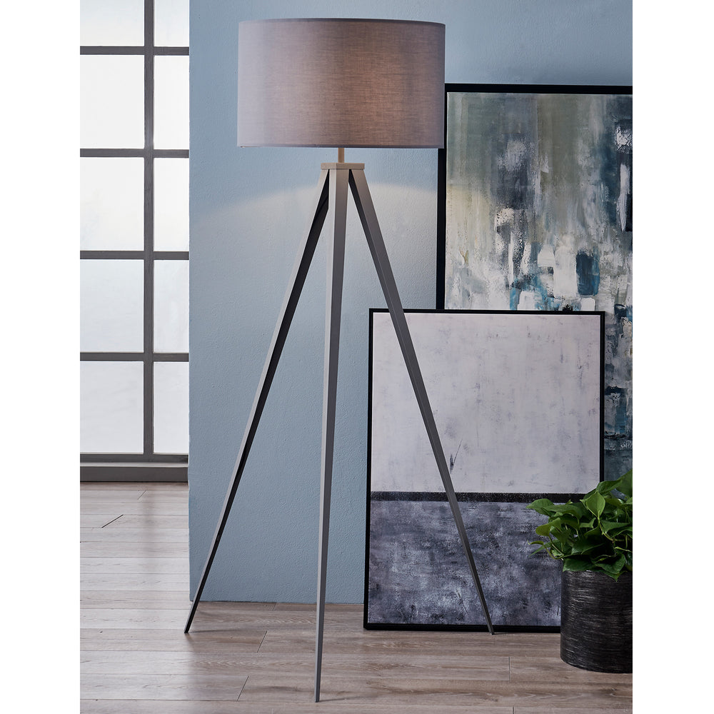 A Teamson Home Romanza 62" Postmodern Tripod Floor Lamp with Drum Shade, Gray stands beside two stacked abstract paintings in a room with hardwood floors.